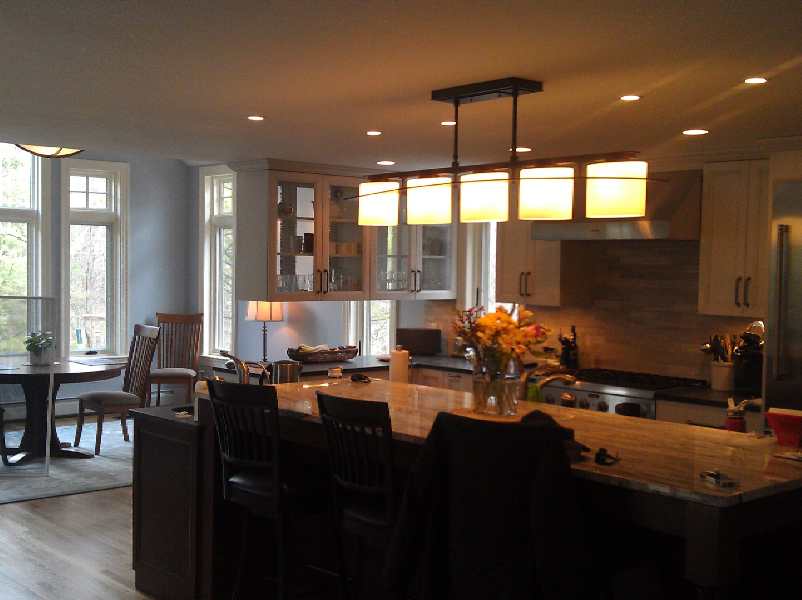 A modern kitchen with a breakfast bar, dark wooden stools, and granite countertops. large windows provide natural light, and there’s a dining area with a table and chairs in the background.