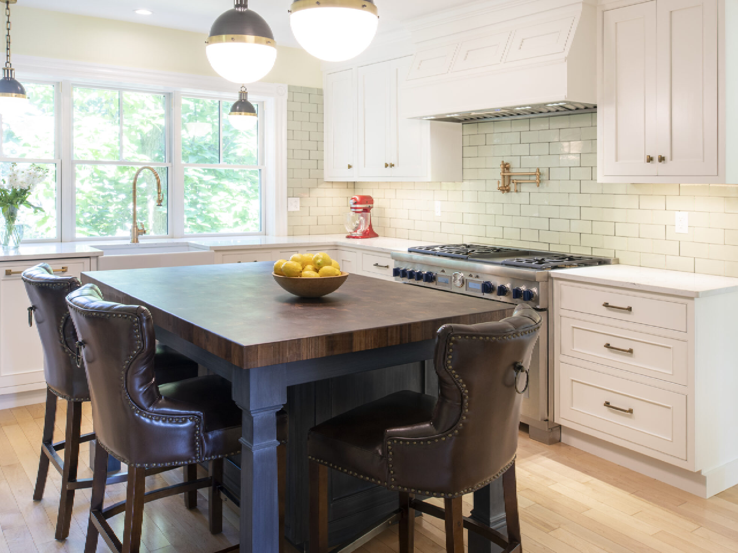 A modern kitchen with white cabinetry, subway tile backsplash, a dark wood kitchen island with brown leather stools, stainless steel appliances, and natural light from large windows.