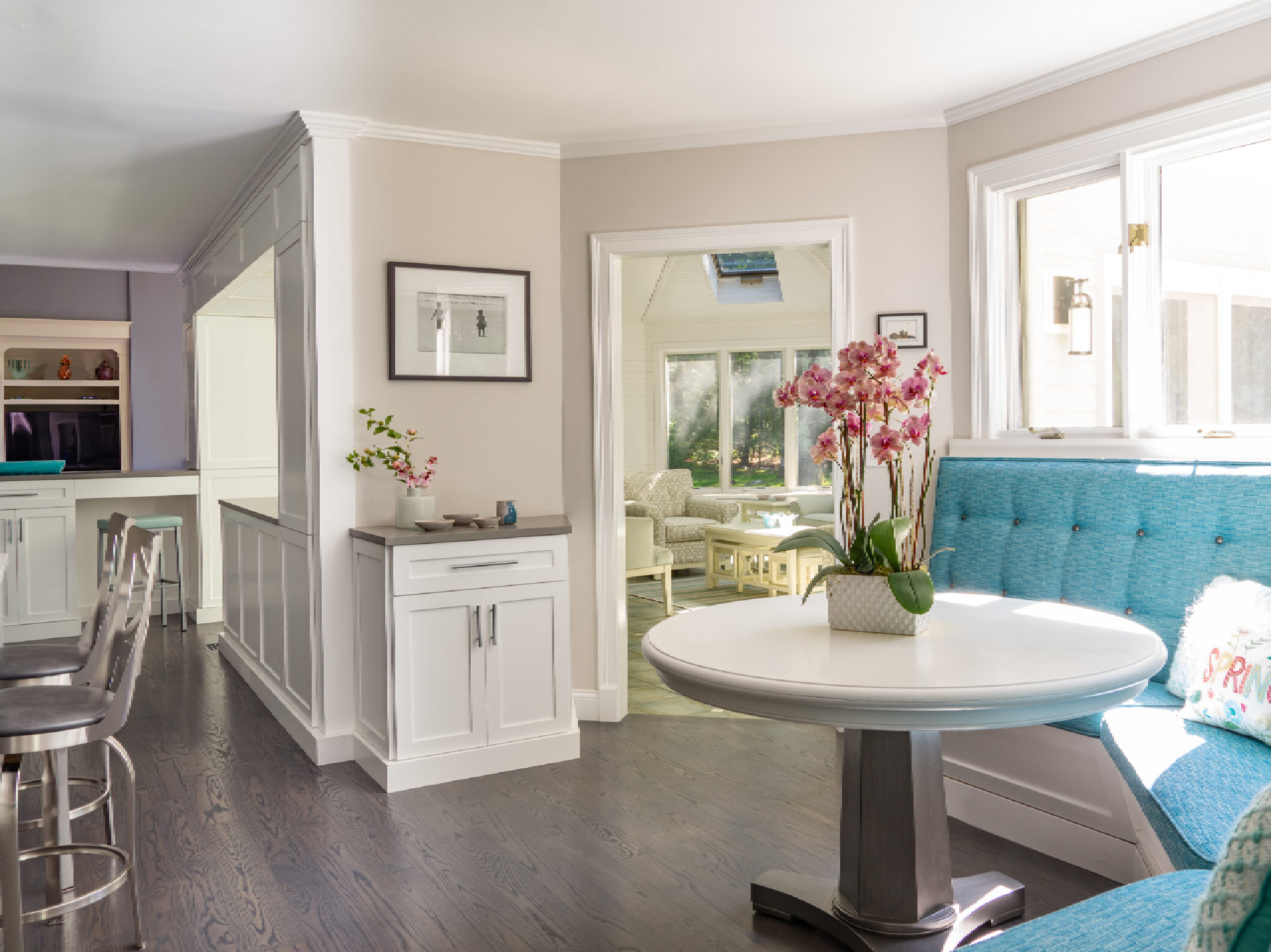 A bright, modern dining area featuring a white round table, turquoise booth seating, bar stools, and elegant dark flooring, with open views into an adjoining room.