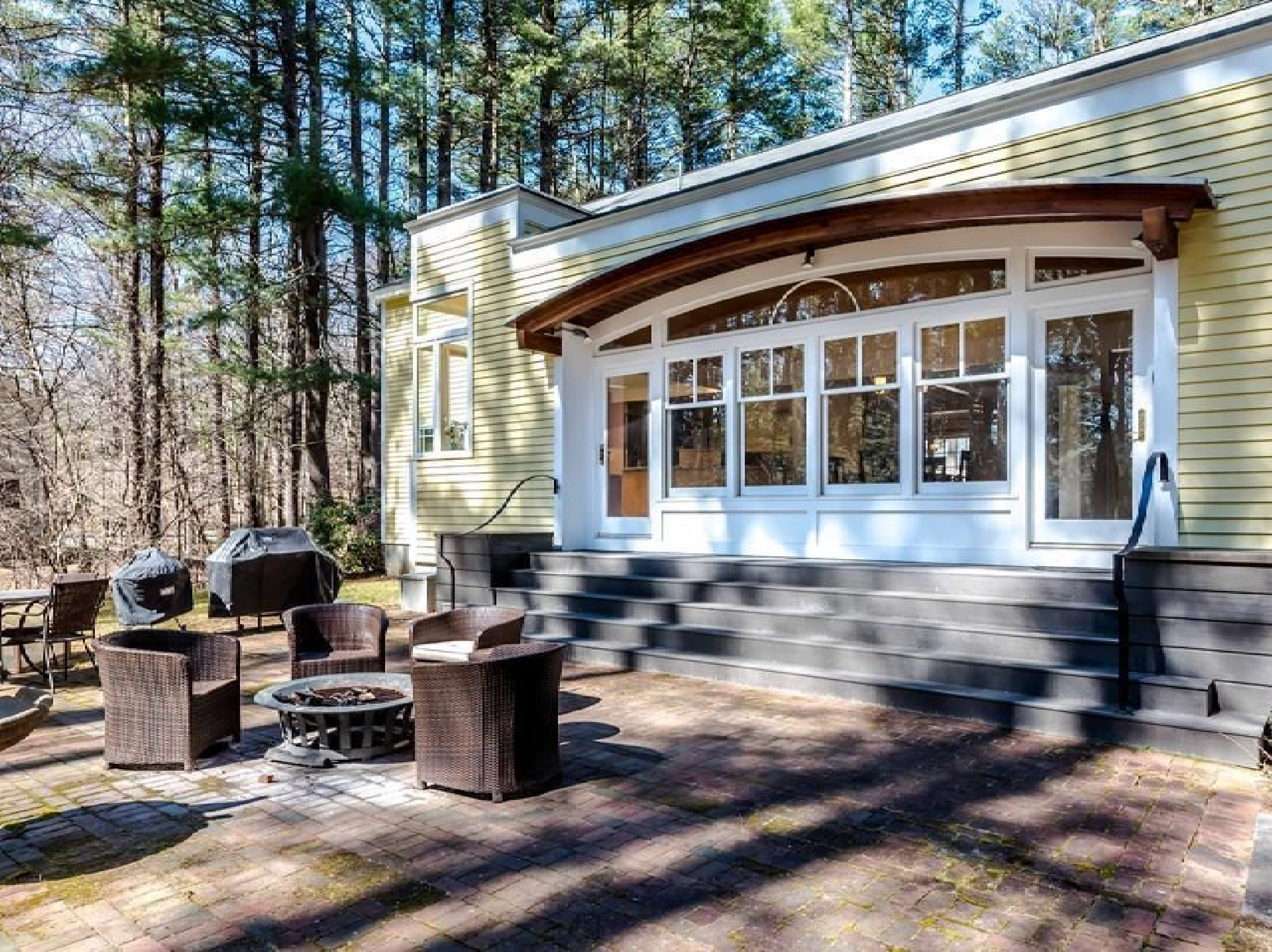 A cozy patio area with woven chairs and a fire pit, set against a curved house with large windows, surrounded by trees under a clear sky.