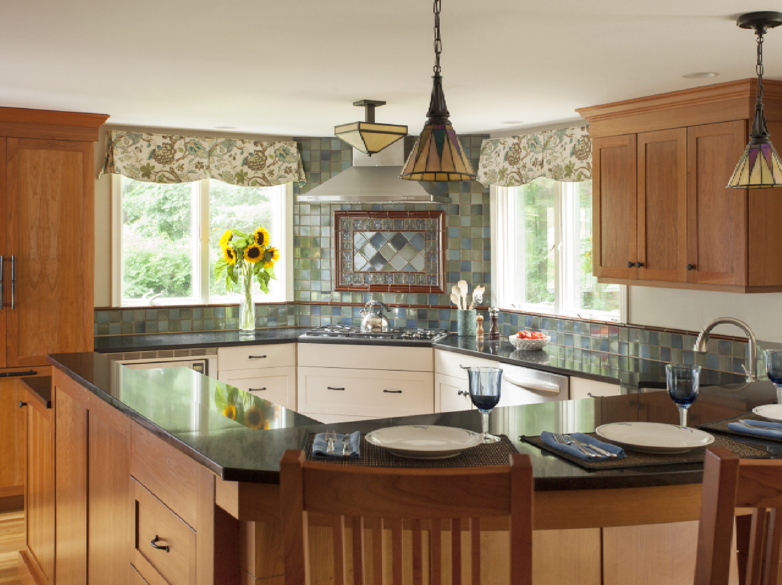 A warmly lit kitchen with wooden cabinets, a central island with a sink, tiled backsplash, and hanging pendant lights. there are place settings on the counter and sunflowers in a vase.